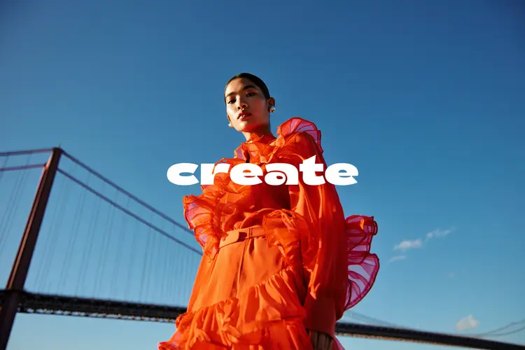 A powerful low-angle fashion portrait of a woman standing confidently in front of a large suspension bridge, shot on a bright, clear day. She wears a dramatic, avant-garde sheer orange ruffled outfit that catches the sunlight and adds rich texture and movement to the image. Her expression is poised and bold, with the shot capturing her from below to emphasize power and presence. The bridge's architectural lines frame her dynamically against a deep blue sky with minimal clouds. The image is captured in 8K resolution with a cinematic color grade — high contrast, slightly warm highlights, crisp shadows, and a soft filmic filter that enhances the drama and editorial quality of the scene. The overall mood is empowering, high-fashion, and editorial-worthy. The word "create" in the the center in a modern, punky font in white.